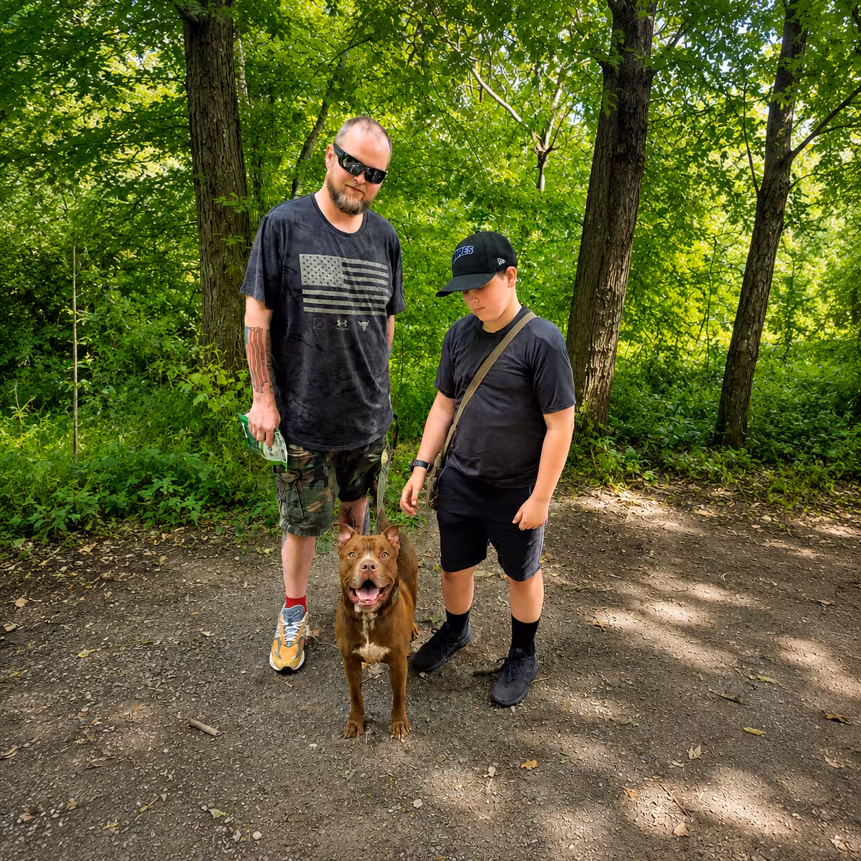 A man and a boy walking a brown pit bull through the woods