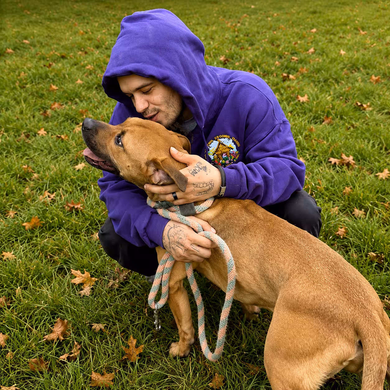 A man in a purple hoodie hugging a tan dog